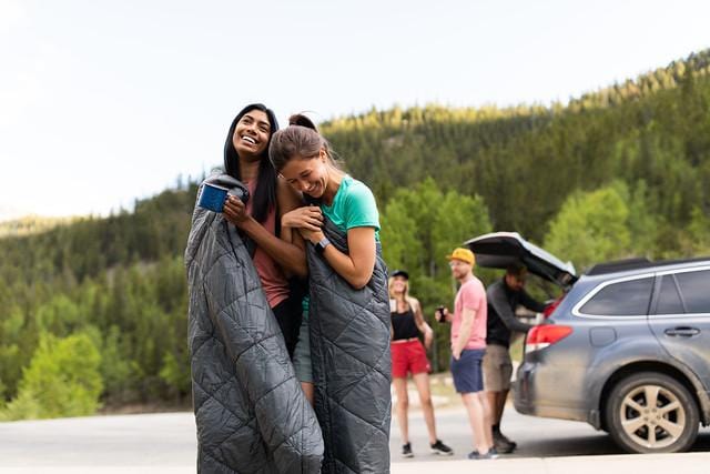GIrls wrapped up in Kammok Quilts smiling in front of a car with people standing behind it i the woods.