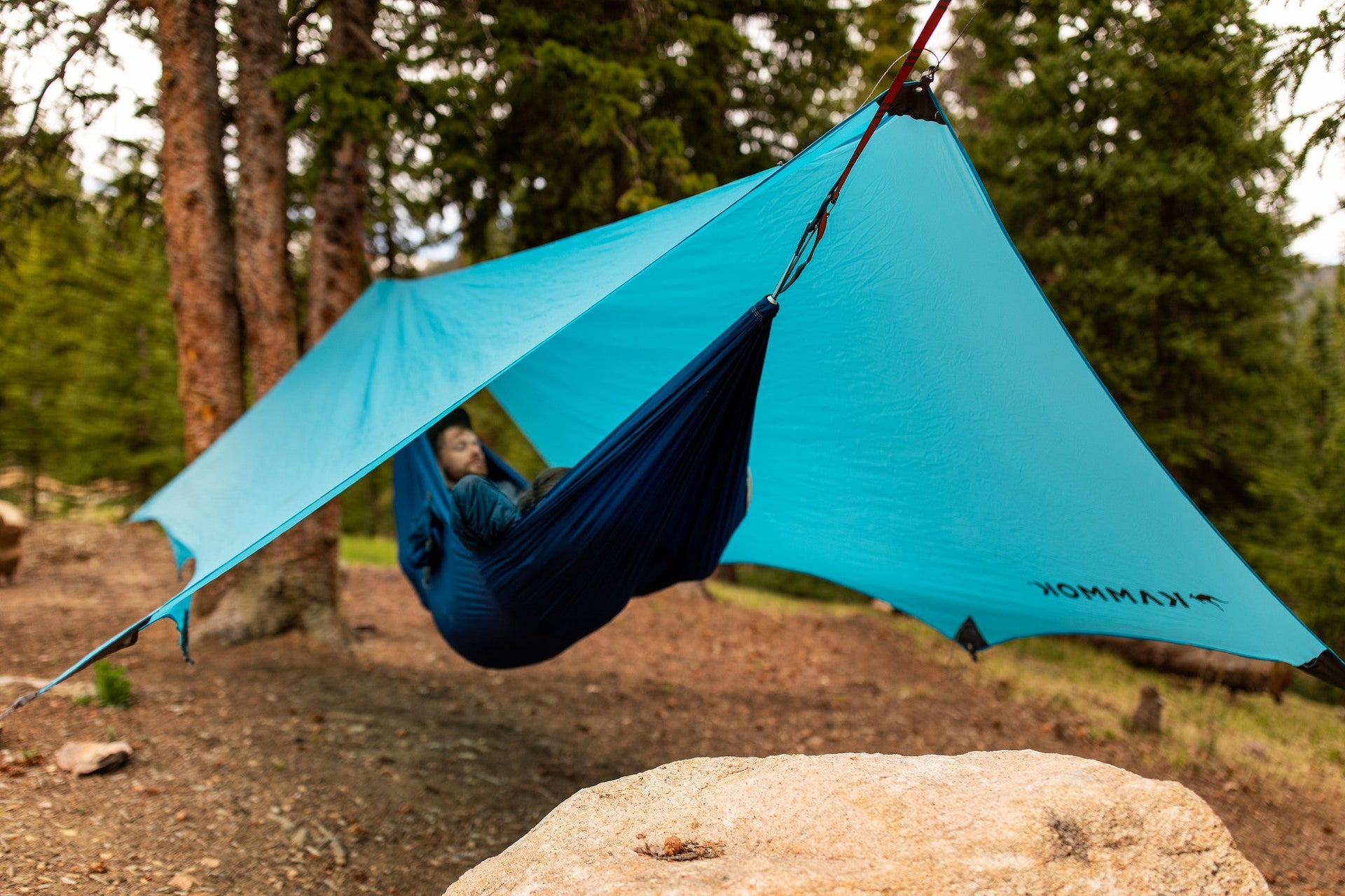 Person resting in a hammock in the forest with a blue Kuhli hammock tarp protecting them from the elements.