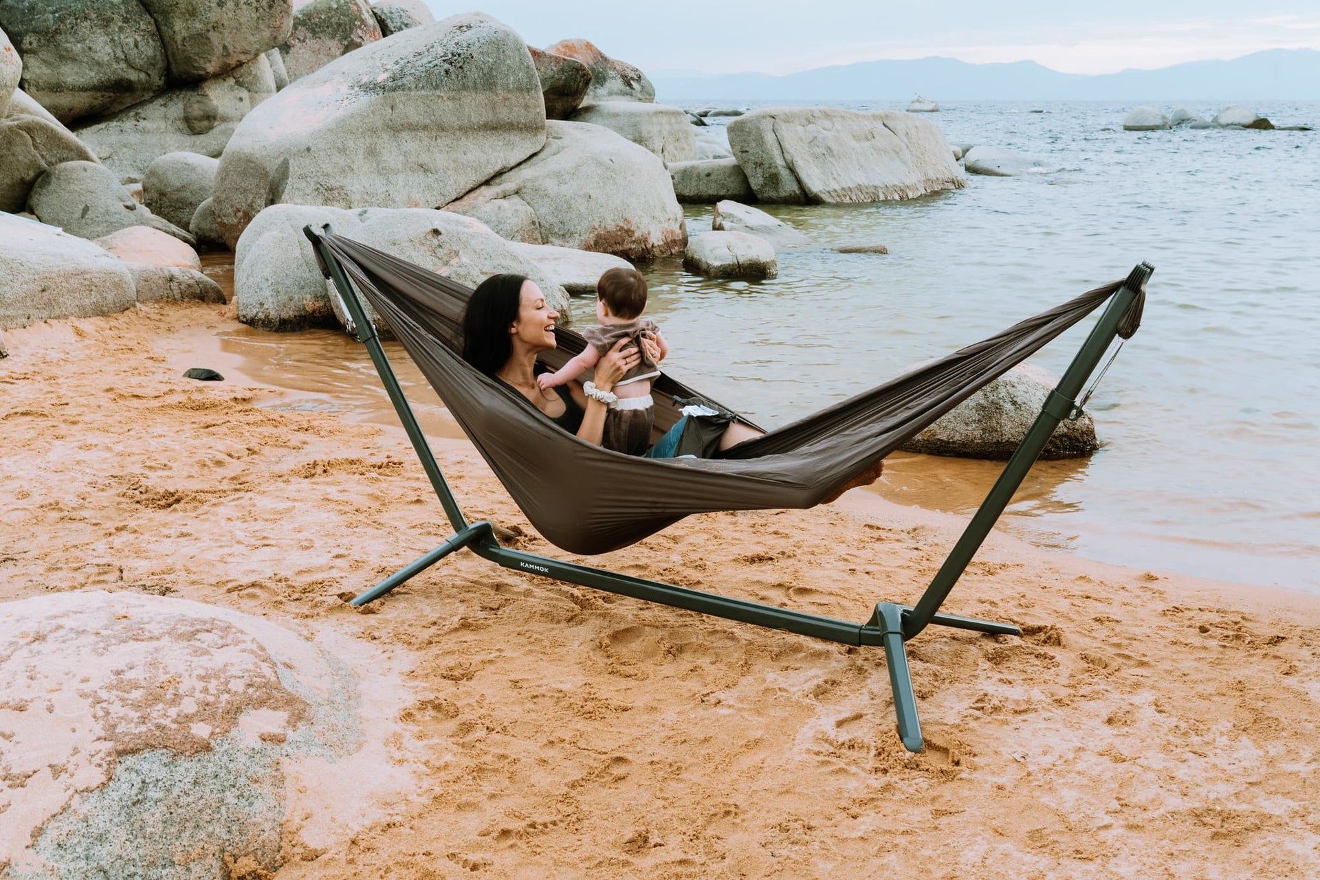 Mom on a rocky beach holding a baby in a Swiftlet Hammock Stand & Gray Hammock.