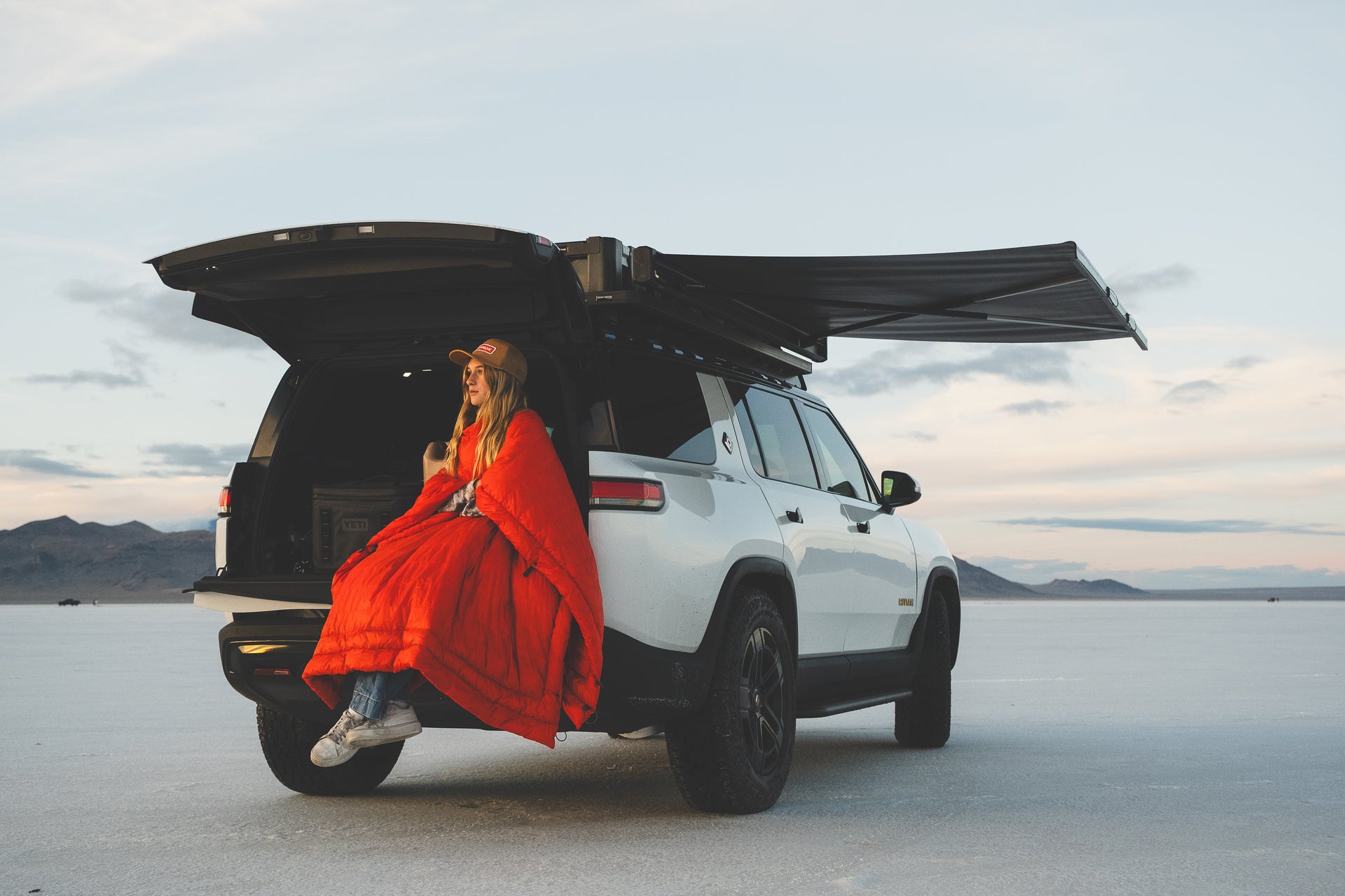 Women sitting wrapped in a Kammok Firebelly Quilt on the trunk of her car overlooking the scenery. The car has a Crosswing Shade Awning extended.