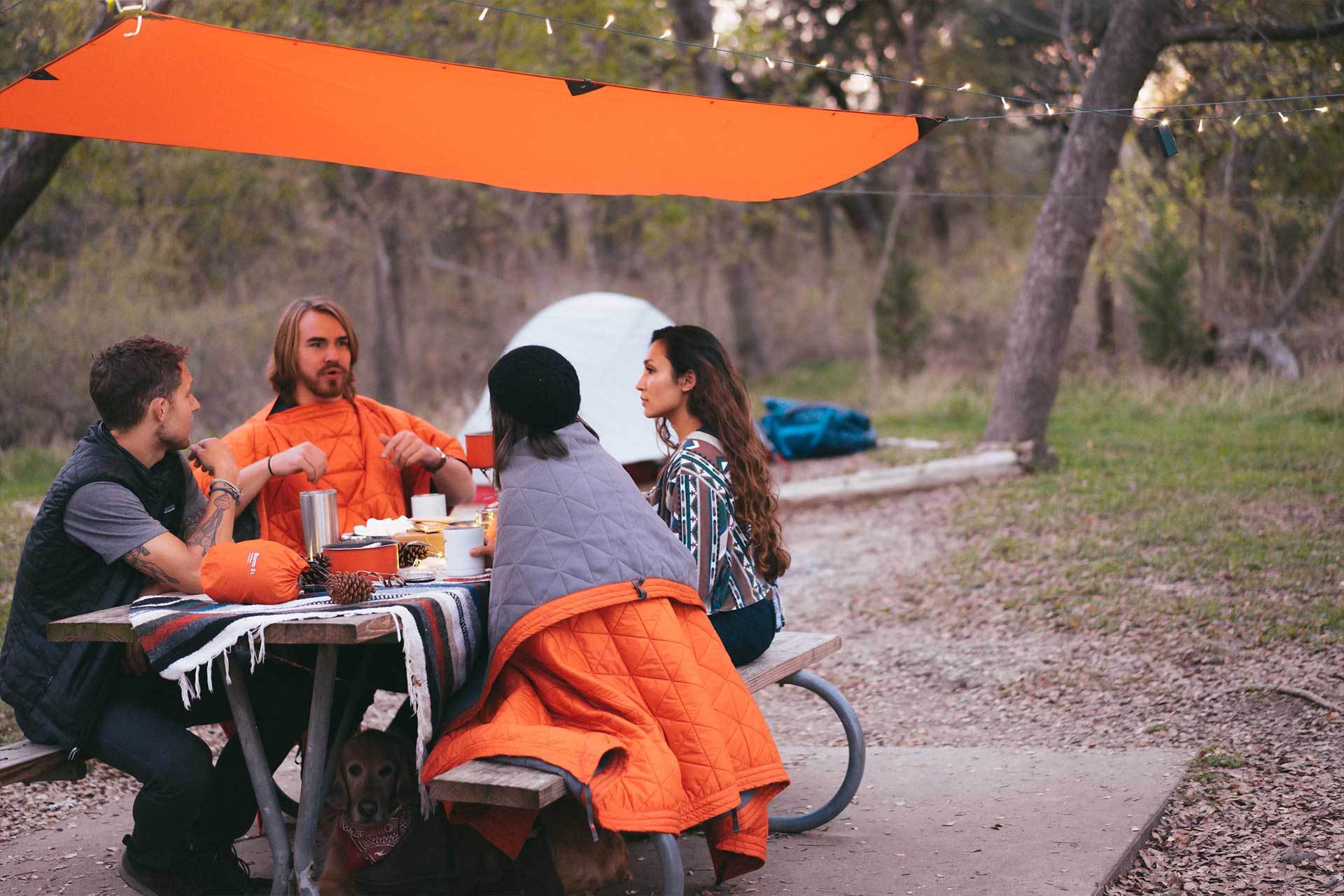 Four people sitting at a picnic table underneath a Kammok Tarp at a campground.