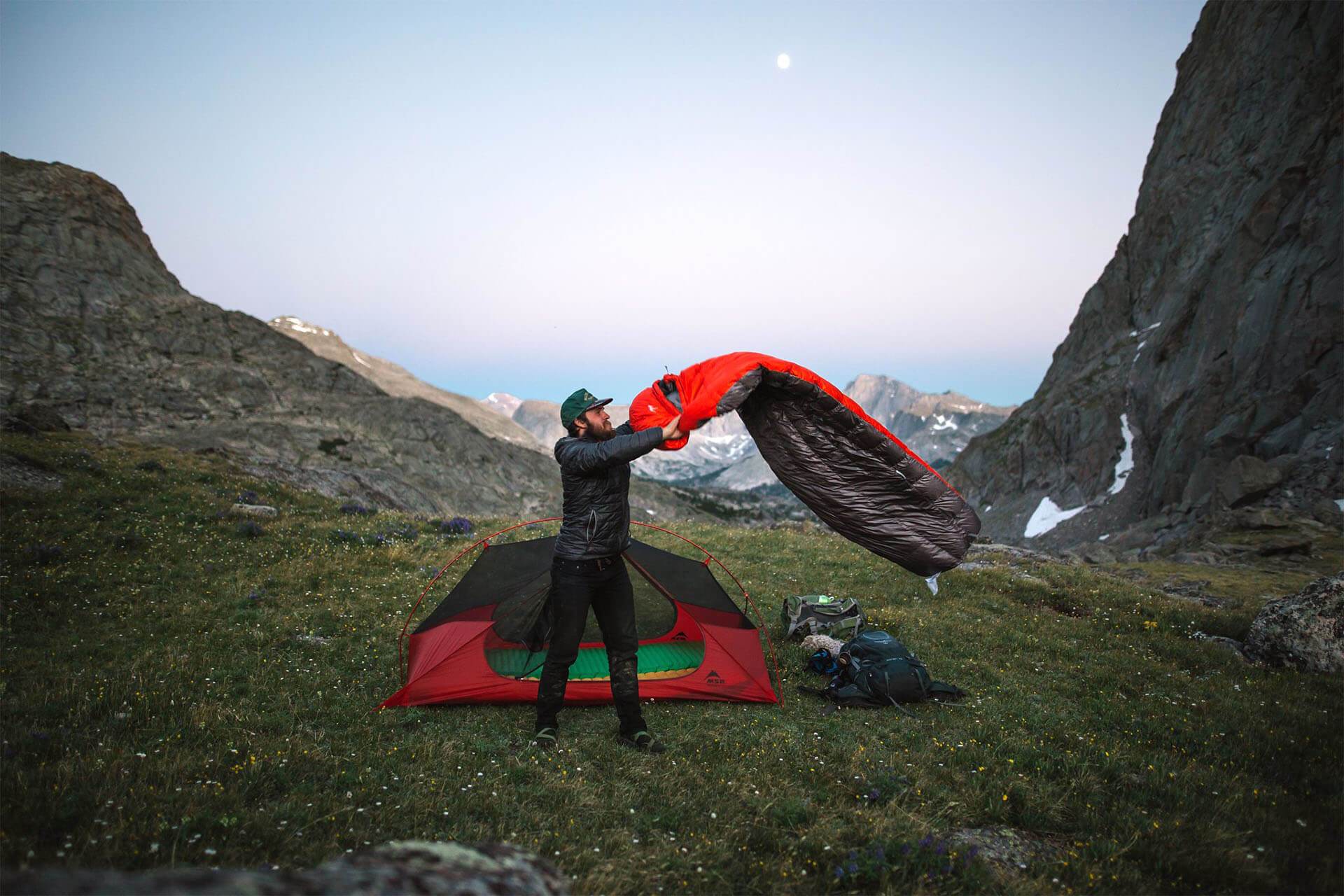 Man shaking out his sleeping bag in front of a tent in a mountain pass.