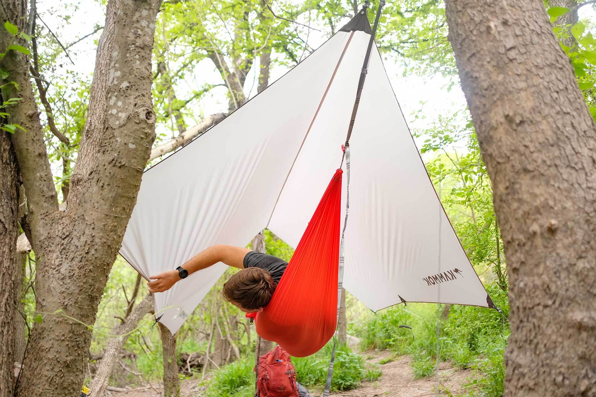A man lying in a hammock tugging on his Kuhli Ultralight Tent Shelter while suspended between two trees.